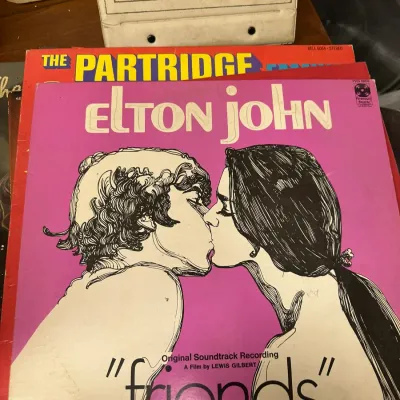 Sale Photo Thumbnail #130: Stack of vintage vinyl LP record albums featuring Elton John's 1971 soundtrack for "Friends" on top, alongside a Partridge Family record and a vintage record storage case.