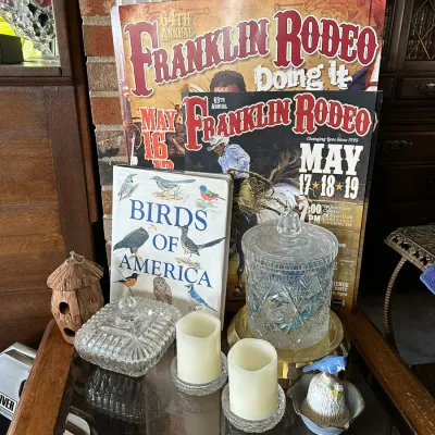 Sale Photo Thumbnail #250: Collection of home decor and vintage memorabilia, featuring Franklin Rodeo posters, a 'Birds of America' book, cut glass covered jars, pillar candles, and a decorative bird figurine.