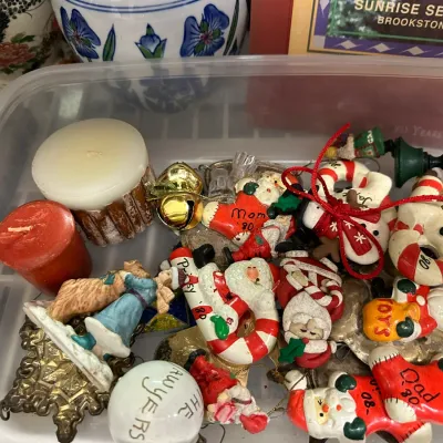 Sale Photo Thumbnail #74: A plastic bin filled with vintage hand-painted Christmas ornaments from around 1980, featuring Santas, snowmen, and personalized candy canes, alongside small decorative candles.