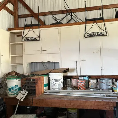 Sale Photo Thumbnail #468: Workshop workbench topped with various supplies including joint compound buckets, paint cans, metal pipes, and wooden organizers, below mounted white storage cabinets.