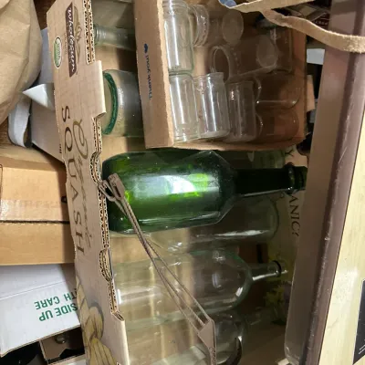 Sale Photo Thumbnail #324: Cardboard box containing an assortment of vintage glass bottles and small jars, including green wine bottles.