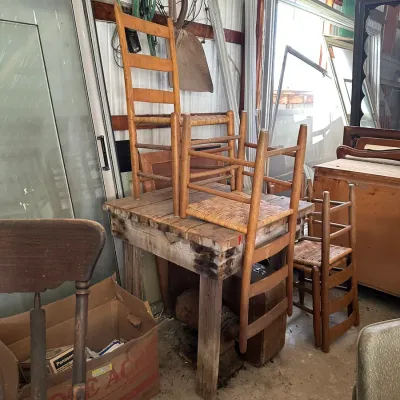 Sale Photo Thumbnail #723: Collection of rustic ladderback chairs with woven seats, stacked on and around a heavy wood workbench in a workshop setting.