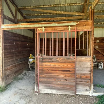 Sale Photo Thumbnail #266: Rustic wooden horse stall featuring a sliding metal frame door with vertical bars and lower wood planking.