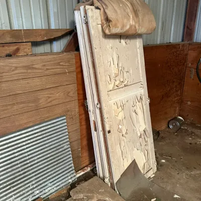 Sale Photo Thumbnail #552: Stack of vintage white-painted wooden doors featuring heavy paint peeling and weathering, stored in a barn with a canvas cover.
