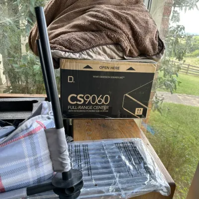 Sale Photo Thumbnail #151: Definitive Technology CS9060 full-range center speaker box sitting on a wooden cabinet, accompanied by a folded fleece blanket and packaged wire racks.
