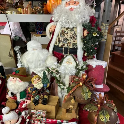 Sale Photo Thumbnail #13: Collection of festive Christmas decorations, including various Santa Claus figurines, oversized metal sleigh bells with ribbon accents, and seasonal floral arrangements.