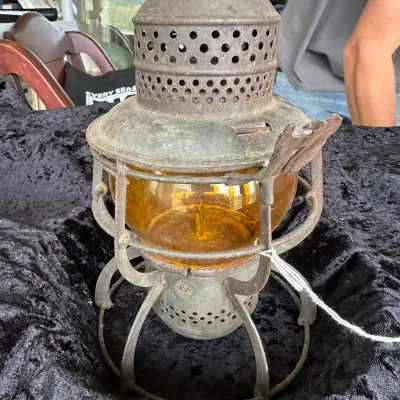 Sale Photo Thumbnail #226: Antique metal railroad lantern featuring a heavy wire cage and an amber glass globe, showing a weathered patina from age and use.