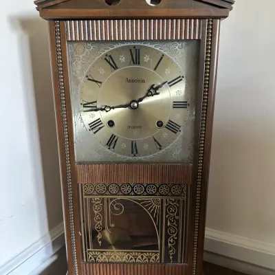 Sale Photo Thumbnail #590: Ansonia 31-day wooden mantle clock featuring Roman numerals, decorative filigree dial detailing, and a gold-tone patterned glass lower door panel.