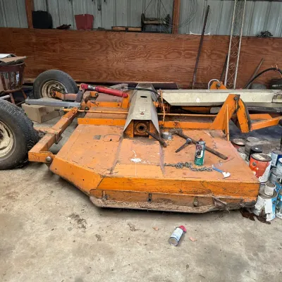 Sale Photo Thumbnail #752: Heavy-duty orange pull-type rotary cutter brush hog featuring dual transport wheels and a hydraulic lift mechanism, stored in a shed.