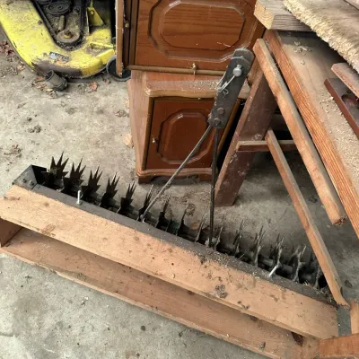 Sale Photo Thumbnail #544: Tow-behind spike lawn aerator attachment with a metal frame and spiked wheels, stored alongside wooden furniture and a riding lawnmower deck.