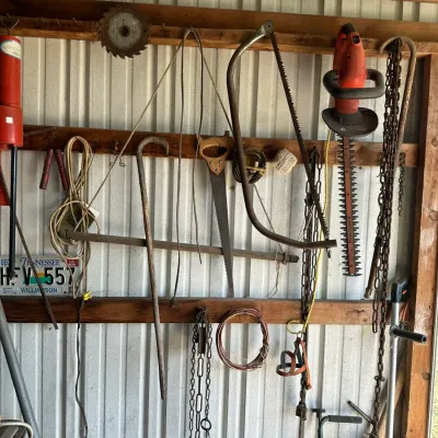 Sale Photo Thumbnail #729: Wall-mounted collection of vintage hand tools and outdoor equipment, including bow saws, chains, a pry bar, and an electric hedge trimmer.