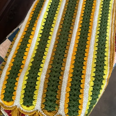 Sale Photo Thumbnail #583: Vintage 1970s crocheted afghan blanket featuring striped patterns in avocado green, gold, yellow, and white yarn, finished with fringed tassels.