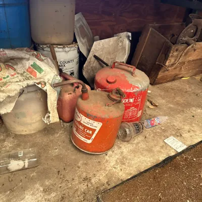 Sale Photo Thumbnail #744: Collection of vintage red metal gas cans and plastic utility buckets stored under a workbench in a workshop or garage setting.