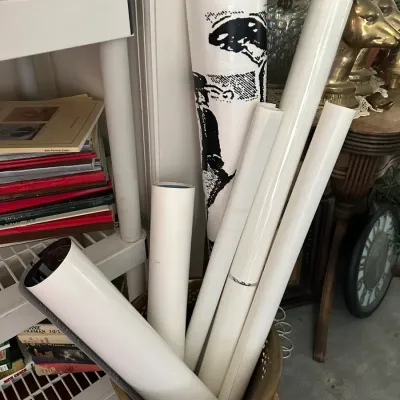 Sale Photo Thumbnail #235: Collection of rolled posters and prints stored in a brass umbrella stand next to shelving with books.