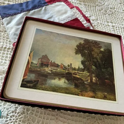 Sale Photo Thumbnail #599: Boxed set of vintage placemats featuring a scenic oil painting print depicting a pastoral landscape with a river and mill.