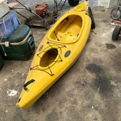 Sale Photo Thumbnail #547: Bright yellow sit-in kayak with front rigging and carrying handles, resting on a concrete floor alongside lawn and garden equipment.