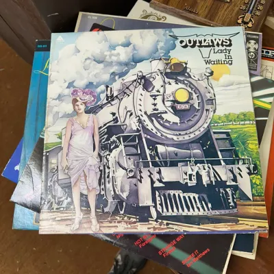 Sale Photo Thumbnail #118: Vinyl LP album cover for Outlaws' 'Lady In Waiting' (1976), resting atop a stack of assorted vintage records and collectibles.