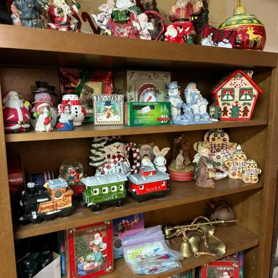 Sale Photo Thumbnail #50: Wooden bookcase filled with various holiday collectibles, including Santa figurines, snowmen, ceramic gingerbread houses, decorative tins, and a ceramic Christmas train set.