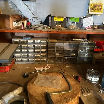 Sale Photo Thumbnail #500: Workshop workbench area featuring vintage parts organizers, hardware boxes, a coping saw resting on a log round, and assorted hand tools.