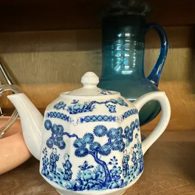 Sale Photo Thumbnail #60: Blue and white porcelain teapot featuring a traditional Asian motif, shown alongside a blue glass pitcher on a wooden shelf.