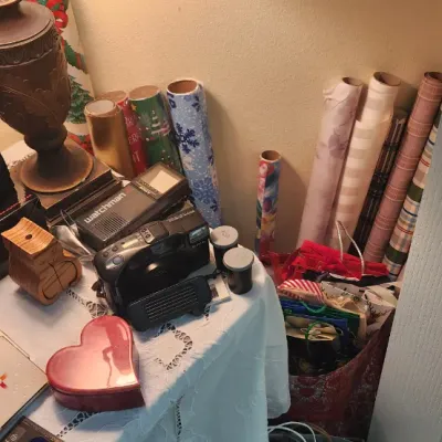 Sale Photo Thumbnail #298: Collection of tabletop items including binoculars, a Sony Watchman portable TV, point-and-shoot camera, decorative wood urn, heart-shaped box, and rolls of wrapping paper.
