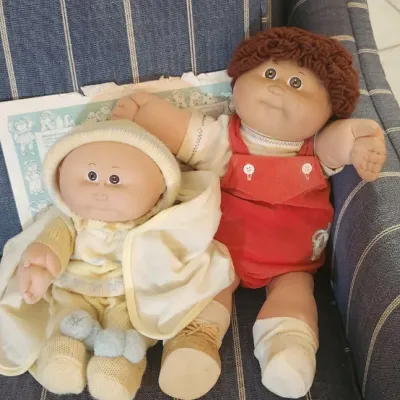 Sale Photo Thumbnail #167: Two vintage 1980s Cabbage Patch Kids dolls, featuring a boy doll in red overalls and a smaller baby doll wrapped in a yellow knit outfit.