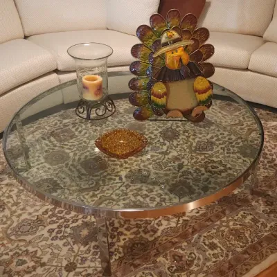 Sale Photo Thumbnail #26: Round glass-top coffee table with a chrome frame, set in front of a large curved off-white sectional sofa with throw pillows.