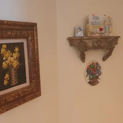 Sale Photo Thumbnail #7: Framed yellow floral oil painting in an ornate gilded frame alongside a wall-mounted carved corbel shelf featuring a Peter Rabbit book and small decor.