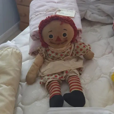 Sale Photo Thumbnail #202: Vintage Raggedy Ann rag doll sitting on a bed among packaged linens and crocheted blankets.