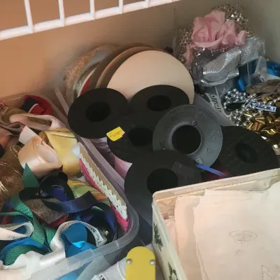 Sale Photo Thumbnail #294: Assorted crafting and sewing supplies, including spools of ribbon, fabric scraps, and trim stored in bins on a closet shelf.