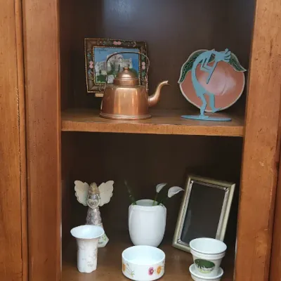 Sale Photo Thumbnail #153: Tall wooden display cabinet filled with decorative home goods including picture frames, ceramics, a copper kettle, and a Kokopelli silhouette.