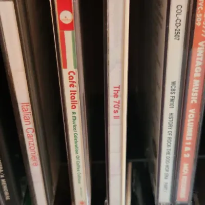 Sale Photo Thumbnail #84: Selection of compact music CDs in jewel cases, including titles such as Cafe Italia, The 70's II, and Glenn Miller, stored in a rack.