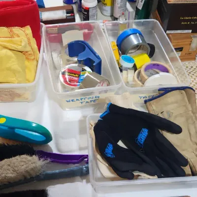 Sale Photo Thumbnail #381: Assorted household utility supplies, including clear storage bins with packing tape, cleaning cloths, work gloves, scrub brushes, and carpet cleaning solution.