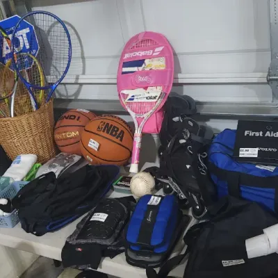 Sale Photo Thumbnail #343: Collection of sporting goods, including tennis racquets, basketballs, rollerblades, golf balls, and a Volvo first aid kit, set on a white folding table in a garage.