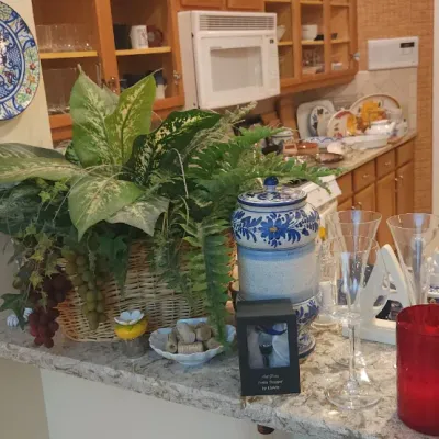 Sale Photo Thumbnail #204: Collection of home decor items including faux plants in a basket, blue and white painted ceramic jar, glassware, and a large decorative bottle displayed on a counter.