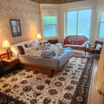 Sale Photo Thumbnail #91: Spacious bedroom featuring a wooden platform bed laden with linens, a wicker settee in the bay window, a large floral patterned area rug, and tables displayed with books and board games.