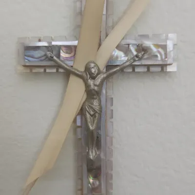 Sale Photo Thumbnail #322: Vintage mother of pearl wall crucifix featuring a metal corpus and abalone shell inlays, wrapped with a cream-colored ribbon.