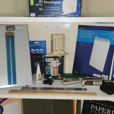 Sale Photo Thumbnail #287: Assorted office and stationery supplies arranged on white shelving, including mailing envelopes, file folders, rulers, staplers, and a Coin Tender counter.