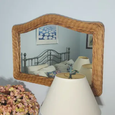 Sale Photo Thumbnail #203: Vintage woven wicker wall mirror featuring a gently arched frame, displayed above a dresser alongside a table lamp and floral arrangement.