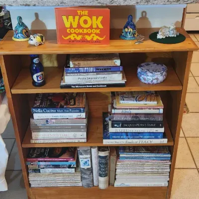 Sale Photo Thumbnail #209: Wooden three-tier bookshelf filled with various cookbooks and kitchen collectibles, including a copy of 'The Wok Cookbook' displayed on top.