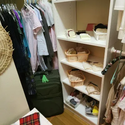 Sale Photo Thumbnail #48: Walk-in closet containing assorted women's clothing, a wide-brim woven straw hat, green rolling luggage, and white shelving units with organized accessories.