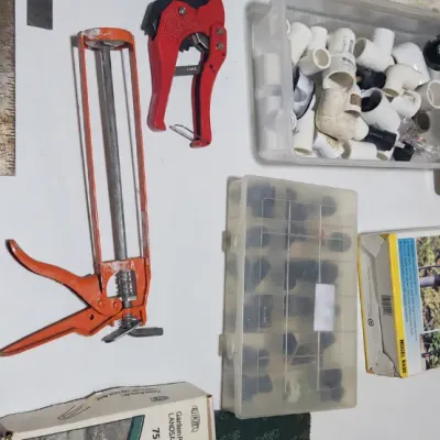 Sale Photo Thumbnail #373: Assortment of hand tools and hardware, including an orange caulk gun, red pipe cutter, L-square, and plastic bins with PVC fittings.