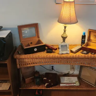 Sale Photo Thumbnail #254: Wicker console table featuring two wooden humidors, a small table lamp, picture frames, and cigar accessories, alongside a wooden stand holding a black HP OfficeJet printer.