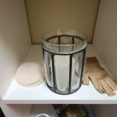 Sale Photo Thumbnail #58: Glass hurricane candle holder with dark metal cage trim, shown on a white shelf alongside a tan glove and fabric pad.