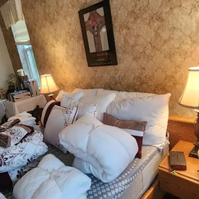 Sale Photo Thumbnail #109: Bedroom set with stacked linens, comforters, and pillows on the bed, alongside an oak nightstand with a traditional table lamp.