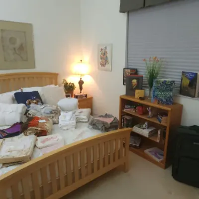 Sale Photo Thumbnail #309: Bedroom set including a light wood bed frame with linens and blankets, a small wooden bookcase, wicker armchair, and various art pieces.