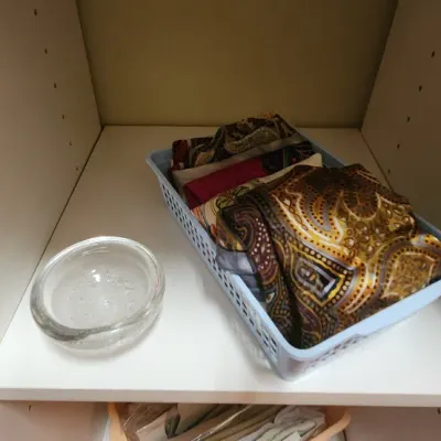 Sale Photo Thumbnail #59: A small clear glass bowl sitting next to a light blue plastic organizer basket filled with folded paisley silk scarves inside a closet shelf.