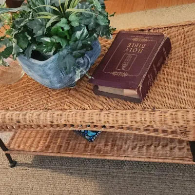 Sale Photo Thumbnail #256: Woven wicker coffee table with a lower storage shelf, displayed with a potted faux plant and a vintage Holy Bible.