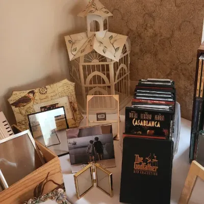 Sale Photo Thumbnail #81: A table arranged with assorted picture frames, DVDs including The Godfather and Casablanca, CDs, and a decorative birdcage.