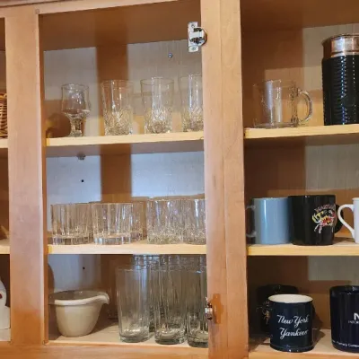 Sale Photo Thumbnail #220: Open kitchen upper cabinets filled with assorted glassware, coffee mugs, a wicker basket, and a Nespresso milk frother.